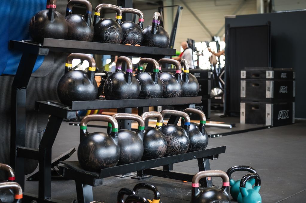 A rack with multiple black kettlebells of varying sizes in a gym setting, with workout equipment in the background.