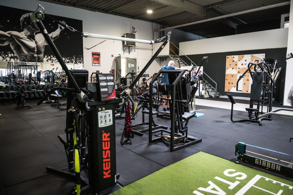 A gym with various exercise machines, a large mural, and a person working out. The floor is covered with mats, and climbing holds are visible on the wall.