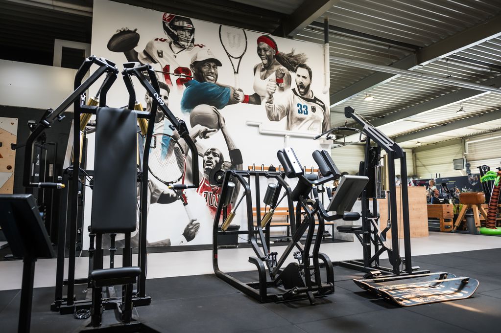Gym interior with exercise machines, under a mural of various athletes from different sports, including football, basketball, and tennis.